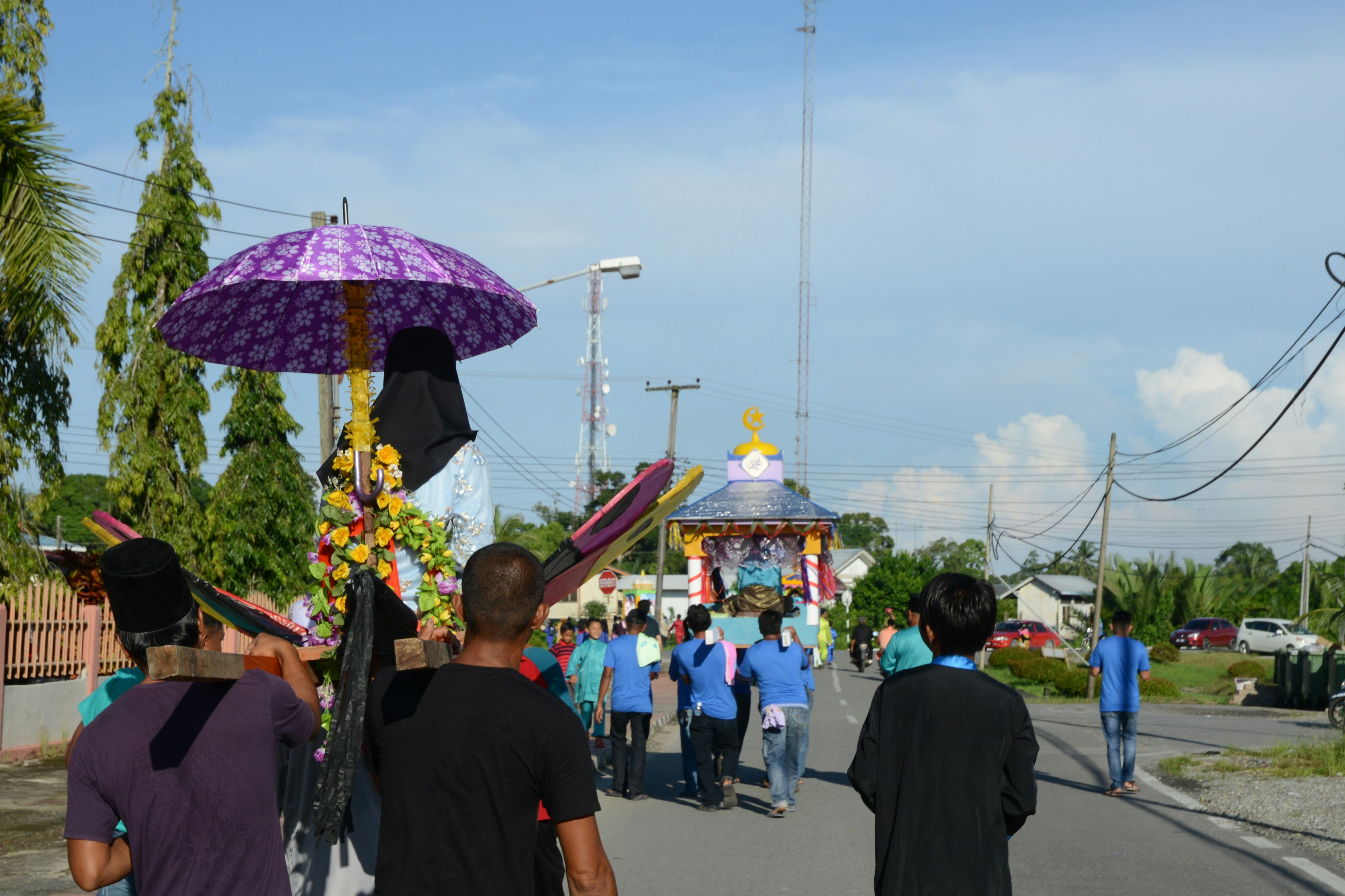A group of people march in the khatamal Al-Quaran parade. There is a figure in an usungan under a purple umbrella.