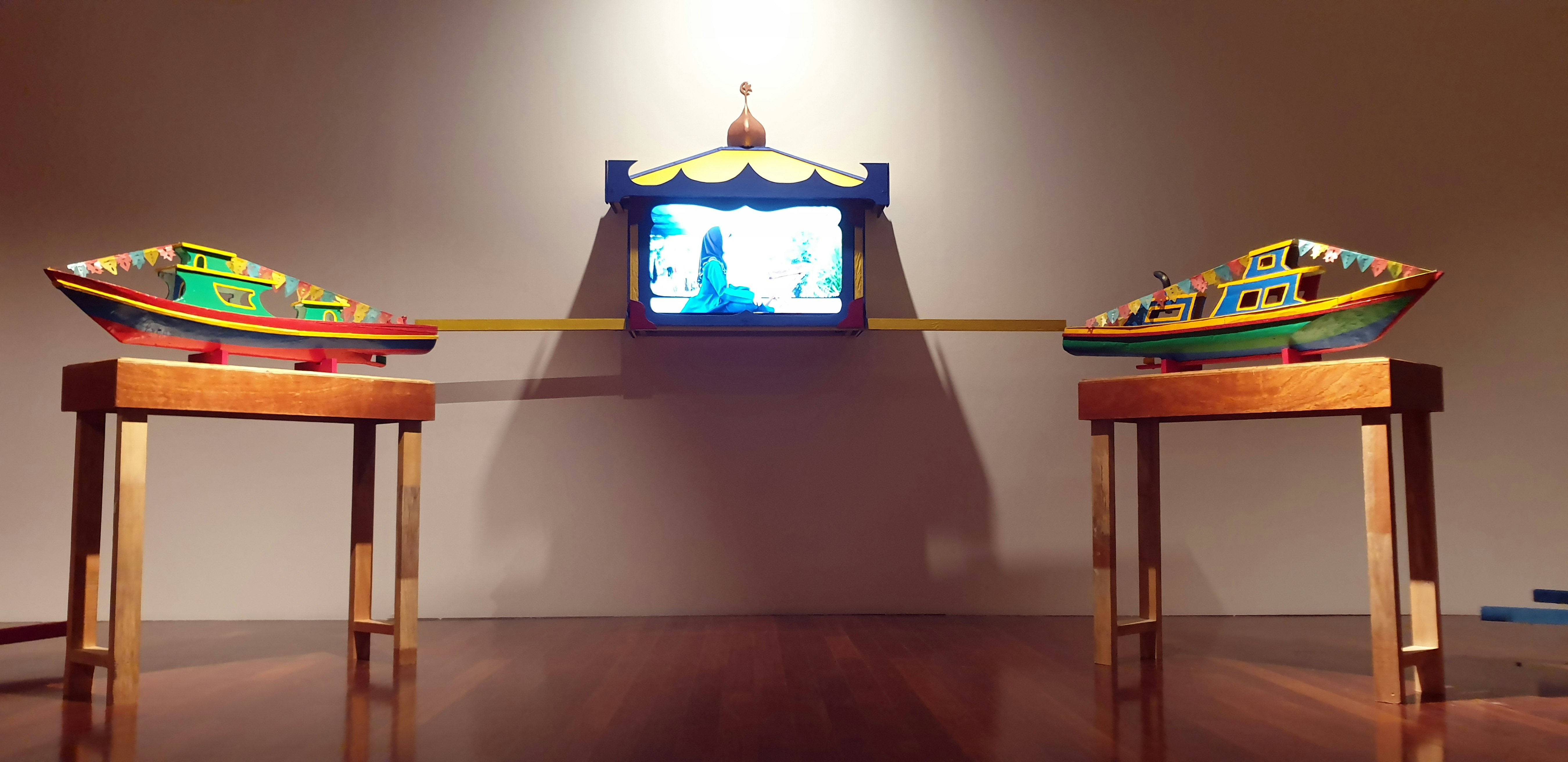 Video artwork nestled between two sculptures resembling usungans placed on wooden tables.