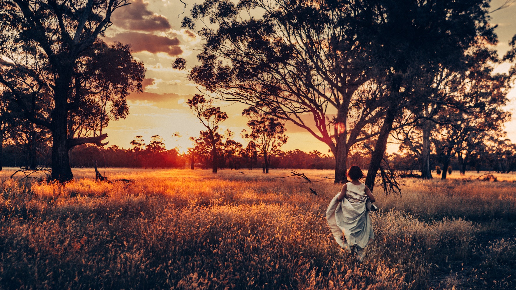 A dancer in a white dress walking through bushland during a sunset