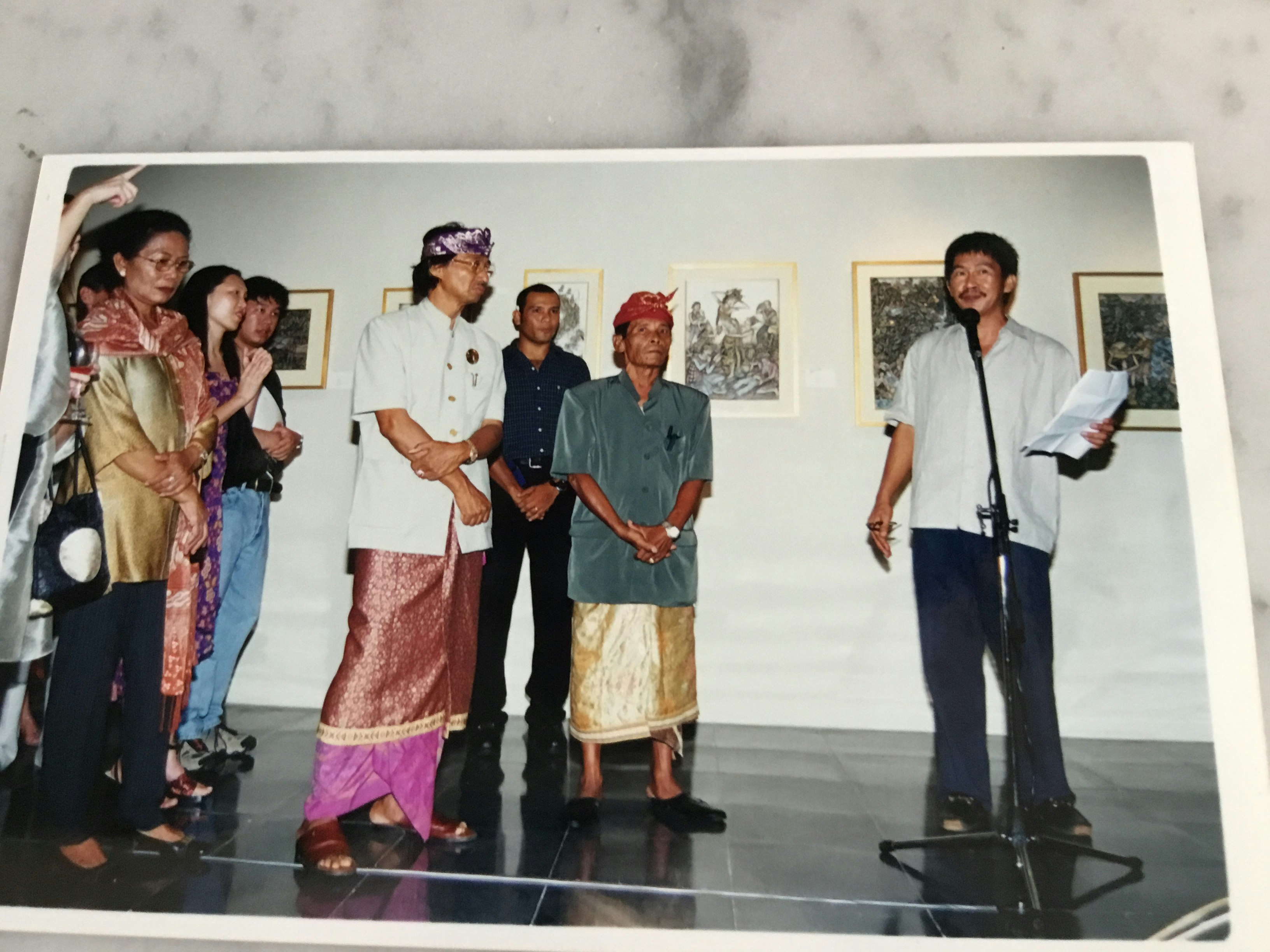 A man in a button up shirt and long navy pants stands at a microphone with a piece of paper in his hand. On his right is a group of people of Southeast Asian appearance, some of whom are wearing batik sarongs and scarves tied around their heads.