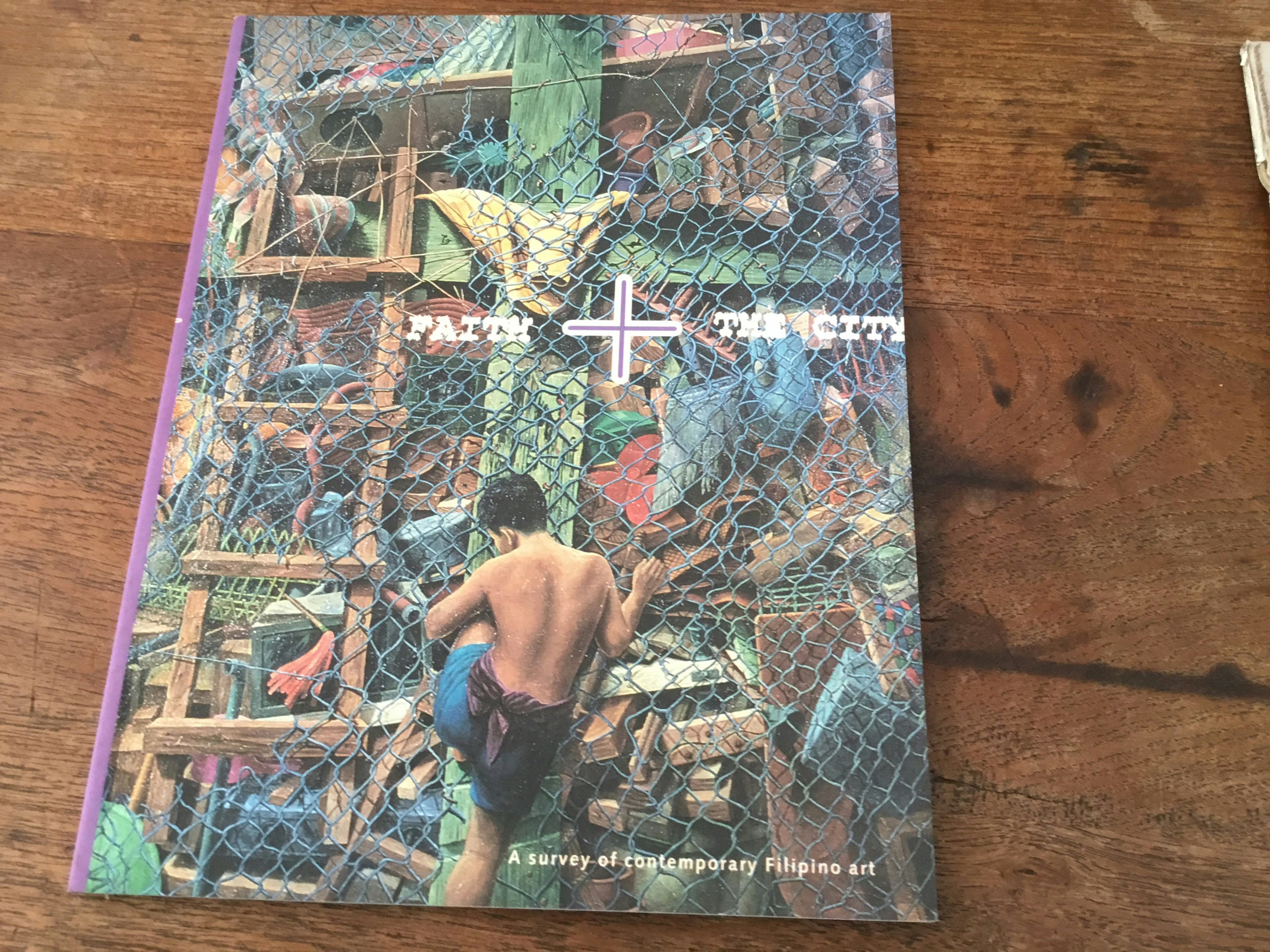 Front cover of a book with a boy climbing a netted fence, title reads 'Faith + The City: A survey of contemporary Filipino art'