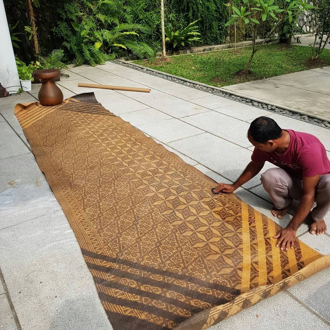 A man squatting on the ground, rubbing a brown woven mat with coconut oil