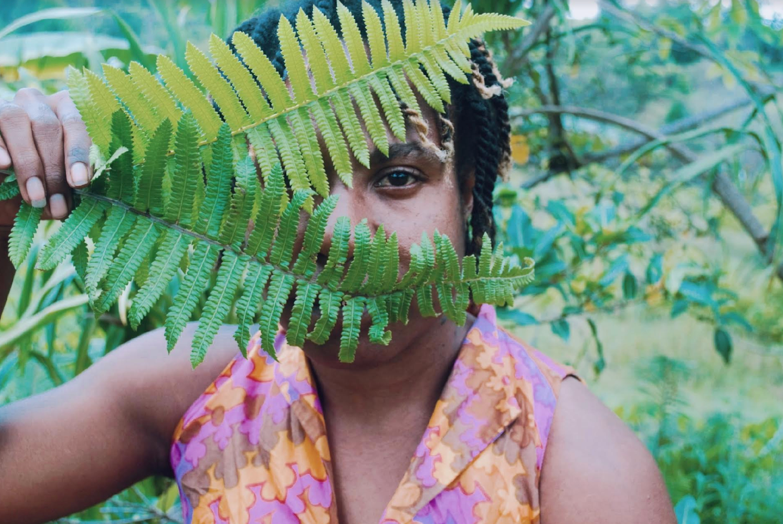 A dark-skinned Polyneasian person with black braids wearing a purple and orange floral vest holds up two green ferns that cover half their face