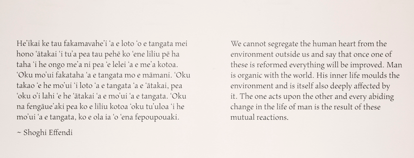 The wall text consists of Tongan on the left and English on the right. Tongan, on the left: He'ikai ke tau fakamavahe'i 'a e loto 'o e tangata mei hono 'ātakai 'i tu'a pea tau pehē ko 'ene liliu pē ha taha ‘i he ongo me'a ni pea 'e lelei 'a e me'a kotoa. 'Oku mo'ui fakataha 'a e tangata mo e māmani. 'Oku takao 'e he mo'ui 'i loto 'a e tangata 'a e 'ātakai, pea 'oku o'i lahi 'e he 'ātakai 'a e mo'ui 'a e tangata. 'Oku na fengāue'aki pea ko e liliu kotoa 'oku tu'uloa 'i he mo'ui 'a e tangata, ko e ola ia 'o 'ena fepoupouaki. English, on the right: We cannot segregate the human heart from the environment outside us and say that once one of these is reformed everything will be improved. Man is organic with the world. His inner life moulds the environment and is itself also deeply affected by it. The one acts upon the other and every abiding change in the life of man is the result of these mutual reactions. – Shoghi Effendi