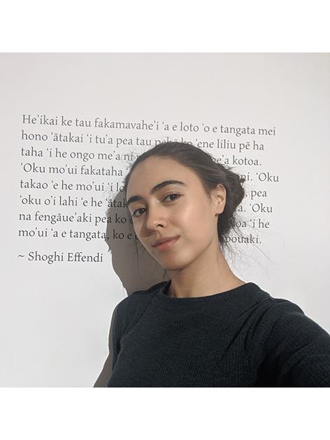 A tanned Pasifika woman in a black top with her black hair in a bun, takes a selfie of her standing against a piece of wall text art. The text is in Tongan, credited to Shoghi Effendi. 