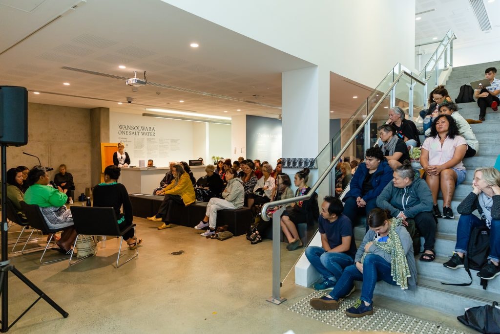 A crowd sitting in an art gallery listening to a group of speakers. Some of the crowd are sitting on the steps listening, while others are seated on some black benches. The decal on the white gallery wall reads: Wansolwara: One Salt Water
