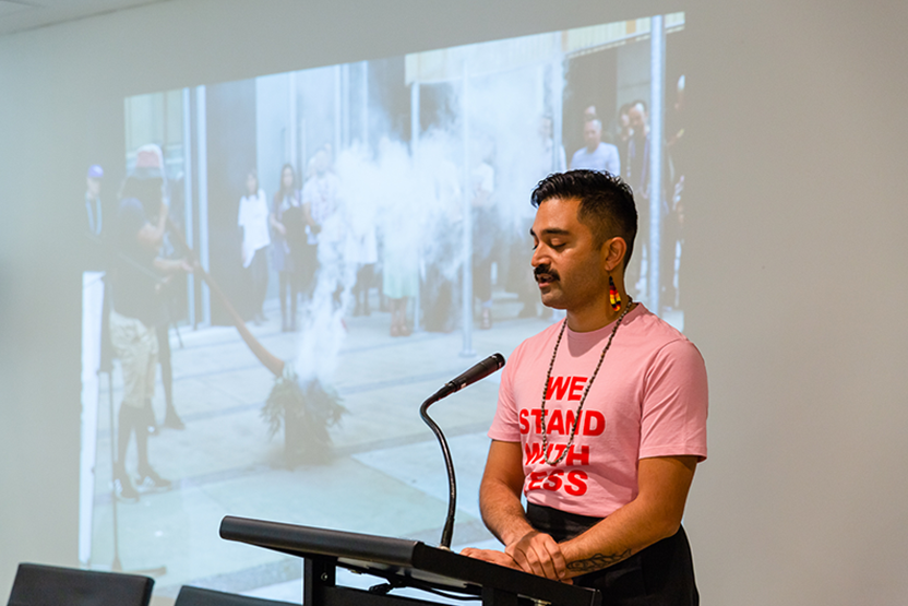 A Pasifika man wearing dangling red, yellow and black earrings and a pink shirt printed with the words 'WE STAND WITH TESS' speaks at a podium while an image of a smoking ceremony is projected behind him. 