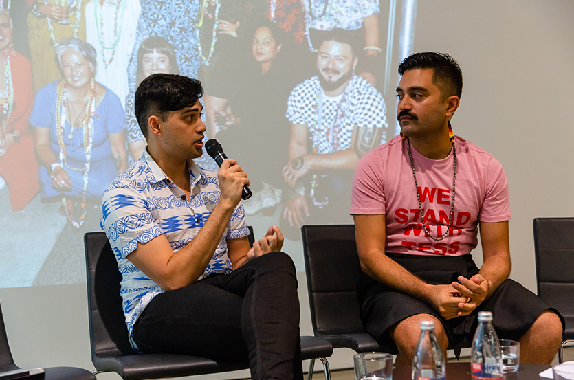 Two Pasifika men sitting on a panel, one (Mitiana Arbon) in a blue geometrically printed button-up shirt and holding a microphone while he speaks to the other (Dr Léuli Eshrāghi), who is wearing dangling earrings, a long necklace and a pink t-shirt printed with the words: WE STAND WITH TESS.