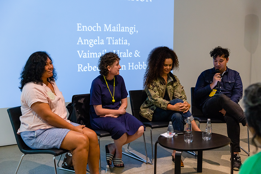 Four Pasifika people sitting on a panel, from left to right: a tanned person with mid-length black curly hair, wearing a pink top and blue striped shorts; a person with curly brown hair, wearing a navy blue shift dress, a bright yellow necklace and black and white slides; a person with long brown curly hair, a camouflage jacket and blue jeans; a person with short black curly hair in a navy blue button up, black pants and sneakers.