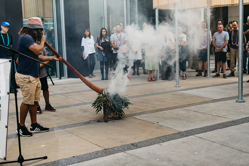 An indigenous person with dreadlocks, wearing a red and black bucket hat, a navy t-shirt, camel brown shorts and sneakers plays a didgeridoo at a smoking ceremony while a crowd watches.