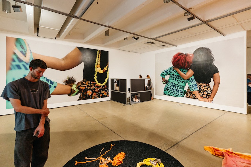 A male-presenting figure stands in an art gallery looking down at a floor installation. Behind him are blown-up photographs of two female-presenting figures with their arms around each other.