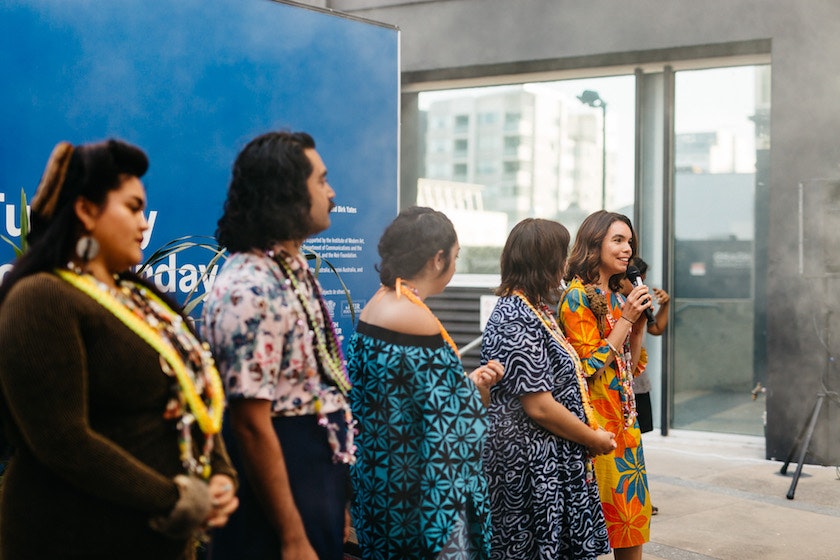 A line of Pasifika people, all in colourful forms of dress, look onto a female-presenting figure in a bright floral dress as she speaks into a microphone.