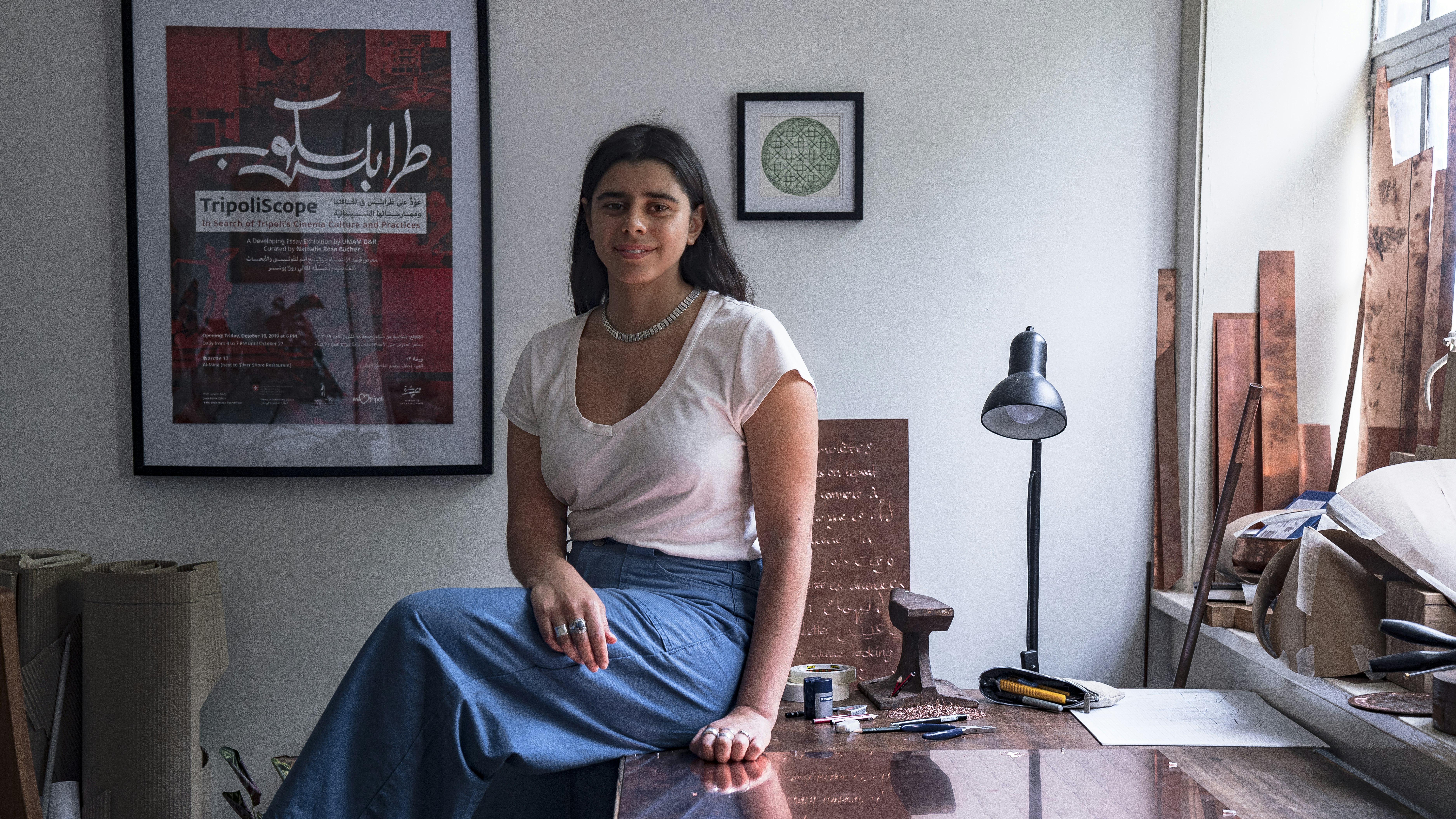 A photo of artist Shireen Taweel sitting on a workbench in her studio.