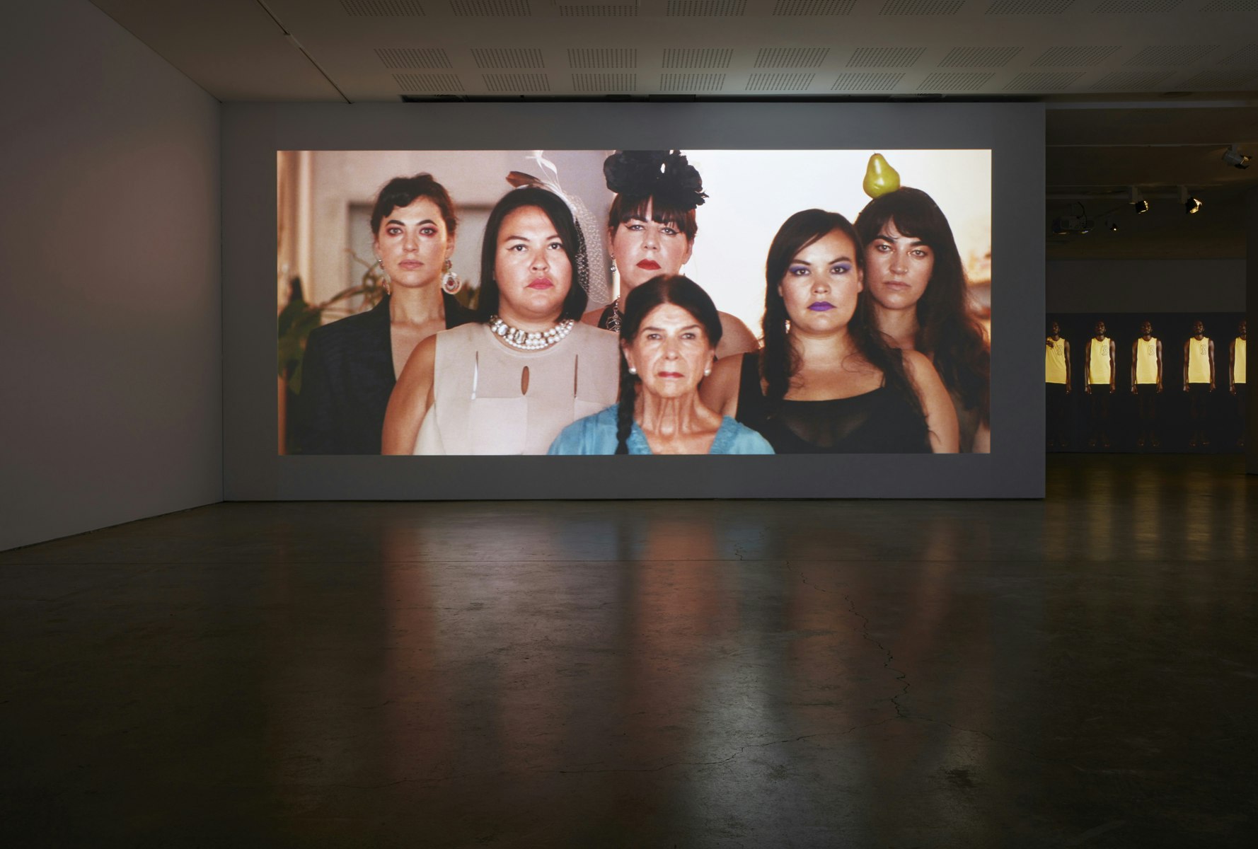 A dark gallery space with a projected still of six Pasifika women facing the photographer.