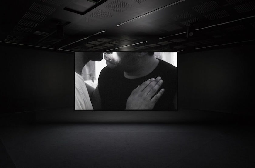 A screen in a dark room, projected with a video still of two figures from the lips down, joined in an embrace.