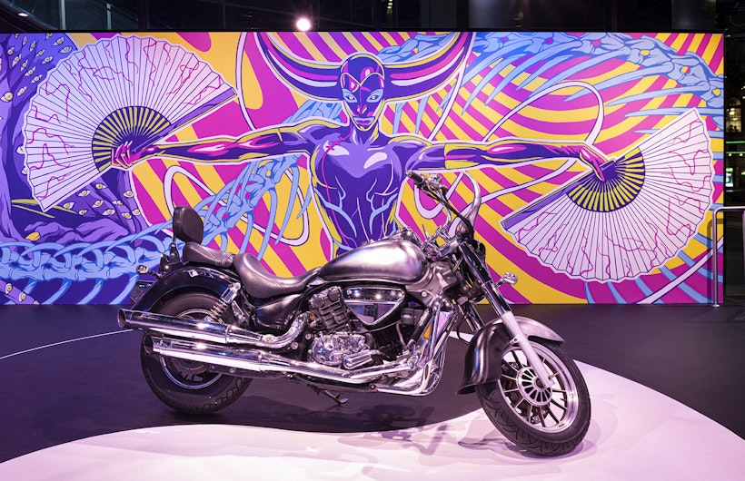 A chrome motorbike is parked in front of a mural of a feminised figure holding her arms out with an opened paper fan in each hand. Two large purple and pink ox horns protrude from her head, while she looks straight ahead with blue ringed eyes and green pupils. Behind her weaves a blue dragon's spine and a series of neon pink and yellow spirals.