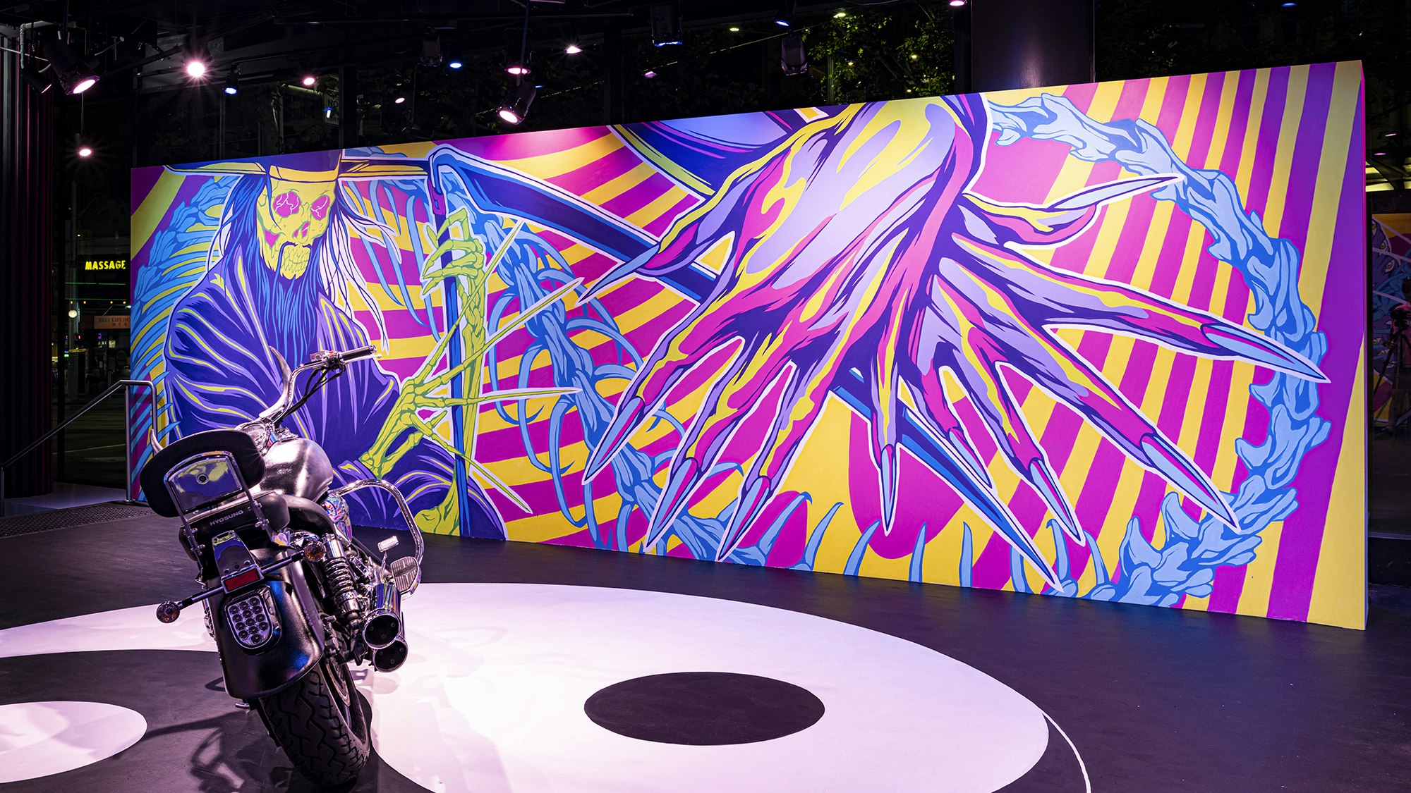 A chrome motorbike is parked in front of a mural with two neon pink, yellow and purple hands crossed over each other. Each hand is the height of the wall and adorned with long pointed fingernails. The hands overlap with a green skeletal Grim Reaper figure in a purple cloak, long blue hair and green skeletal hands holding a purple scythe.