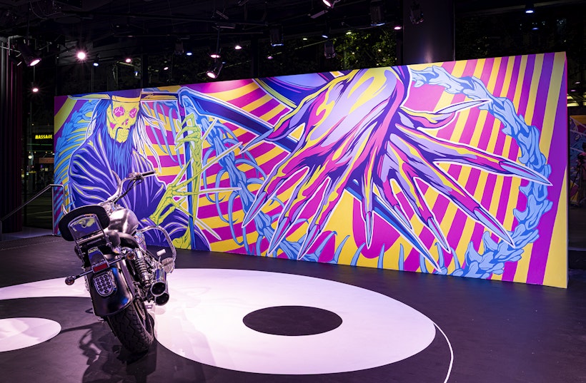 A chrome motorbike is parked in front of a mural with two neon pink, yellow and purple hands crossed over each other. Each hand is the height of the wall and adorned with long pointed fingernails. The hands overlap with a green skeletal Grim Reaper figure in a purple cloak, long blue hair and green skeletal hands holding a purple scythe.