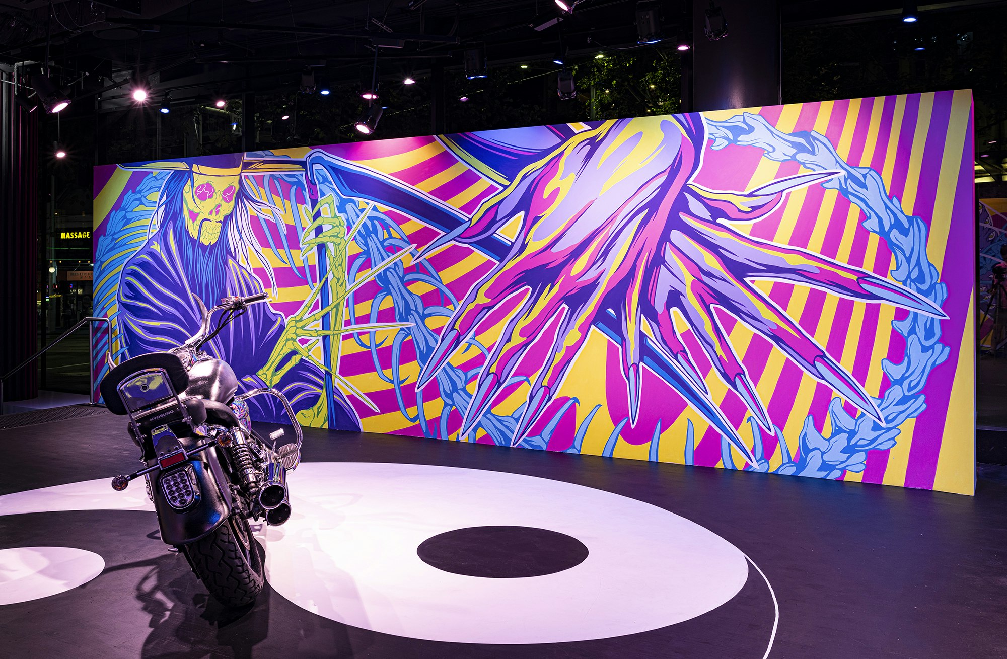 A chrome motorbike is parked in front of a mural with two neon pink, yellow and purple hands crossed over each other. Each hand is the height of the wall and adorned with long pointed fingernails. The hands overlap with a green skeletal Grim Reaper figure in a purple cloak, long blue hair and green skeletal hands holding a purple scythe.