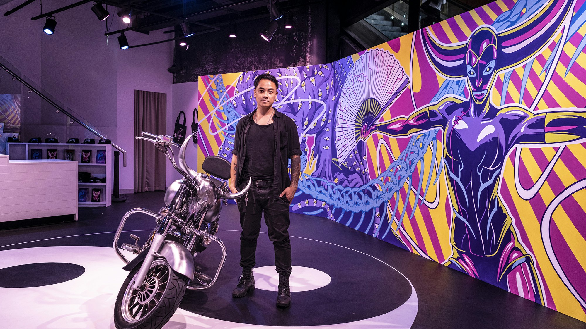 A Thai-Australian artist in a black top and black pants and boots stands next to a chrome motorbike against a long neon pink and yellow mural.