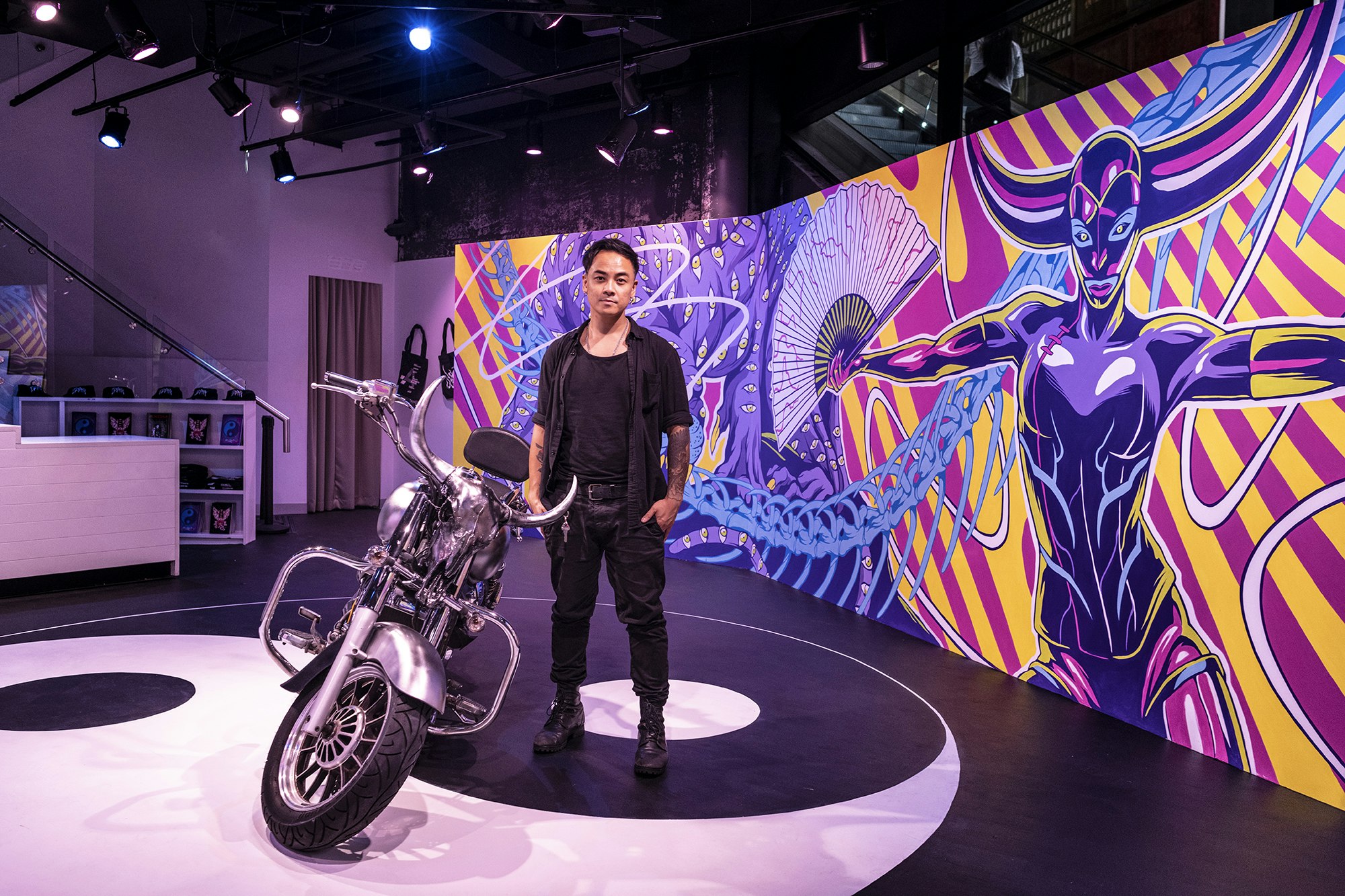 A Thai-Australian artist in a black top and black pants and boots stands next to a chrome motorbike against a long neon pink and yellow mural.