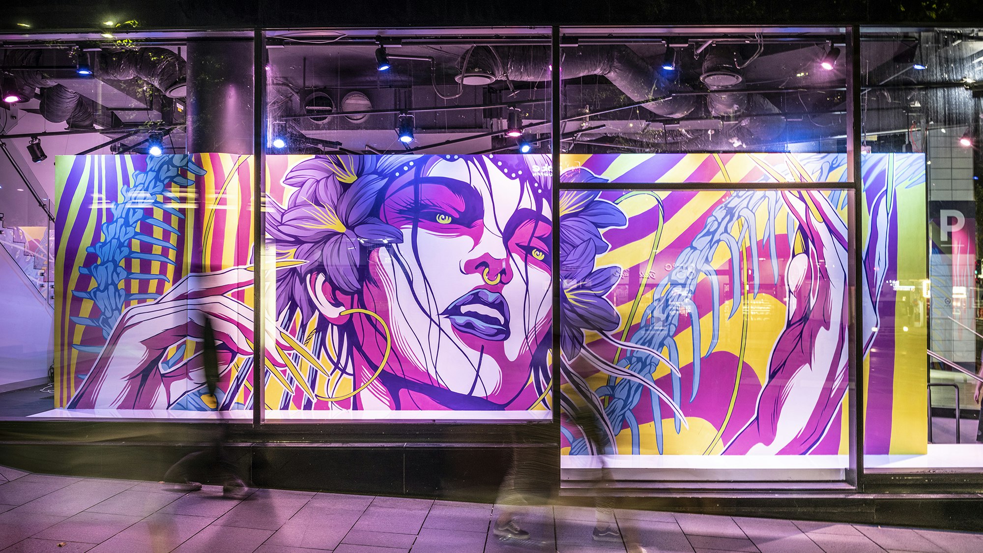 Behind a glass gallery front is a neon pink mural of a woman with purple lilies in her hair, neon pink shadows on the contours of her face, green pupils and blue lips. She has a gold septum ring in her nose. Her hands are raised by her face.