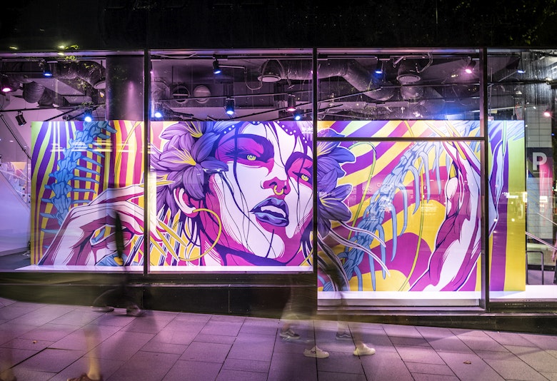 Behind a glass gallery front is a neon pink mural of a woman with purple lilies in her hair, neon pink shadows on the contours of her face, green pupils and blue lips. She has a gold septum ring in her nose. Her hands are raised by her face.