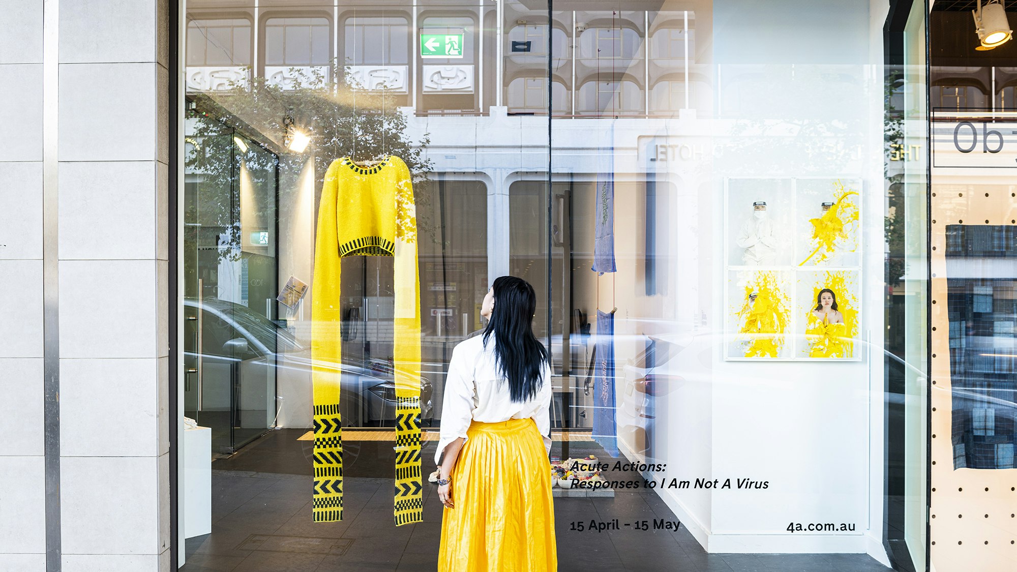 A woman with long black hair in a white blouse and yellow full-length skirt stands on a street outside a gallery glass front, looking at a yellow knitted sweater with sleeves knitted to be 1.5 metres long. The decal sign on the glass front reads 'Acute Actions: Responses to I Am Not A Virus. 15 April - 15 May'