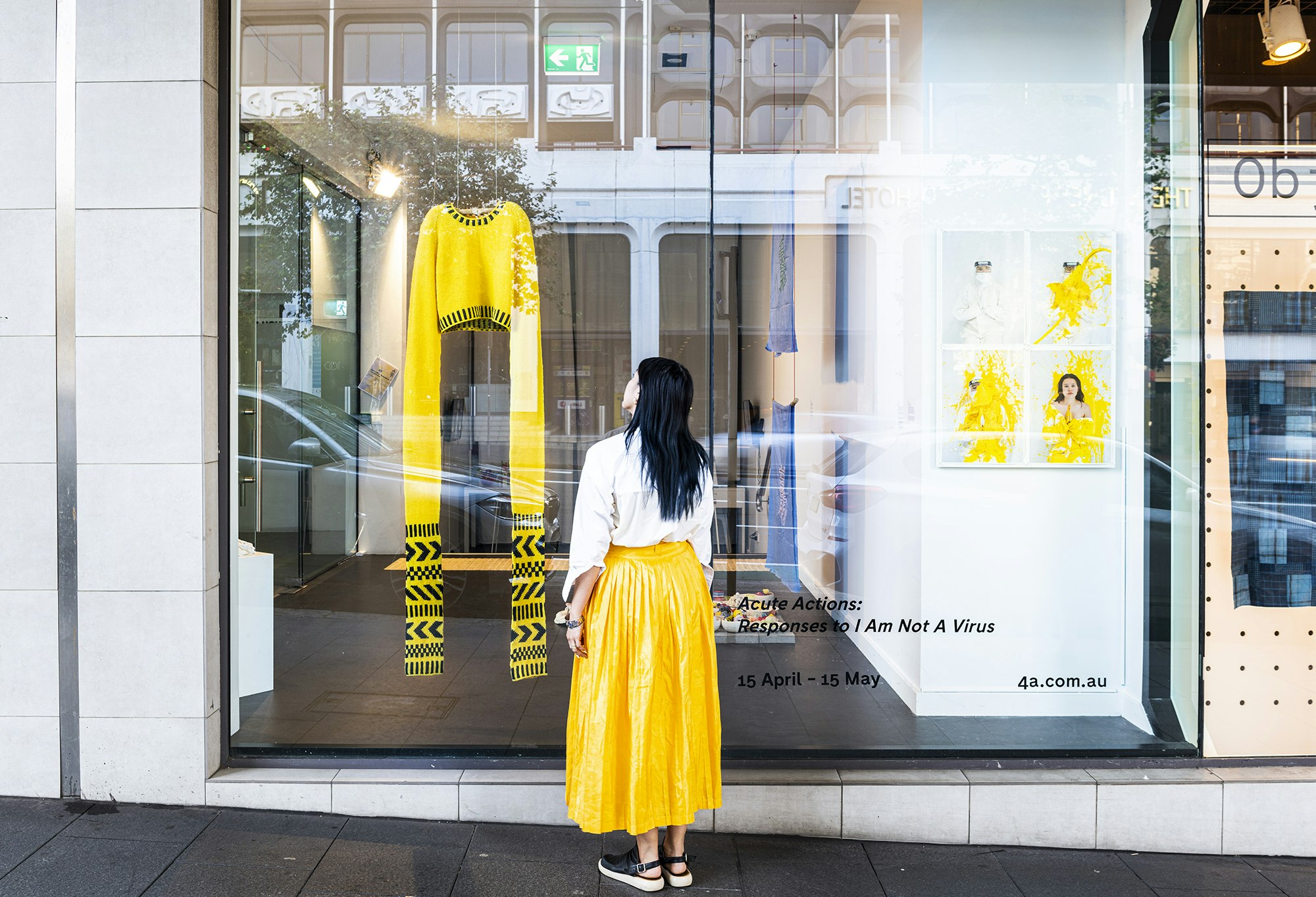 A woman with long black hair in a white blouse and yellow full-length skirt stands on a street outside a gallery glass front, looking at a yellow knitted sweater with sleeves knitted to be 1.5 metres long. The decal sign on the glass front reads 'Acute Actions: Responses to I Am Not A Virus. 15 April - 15 May'