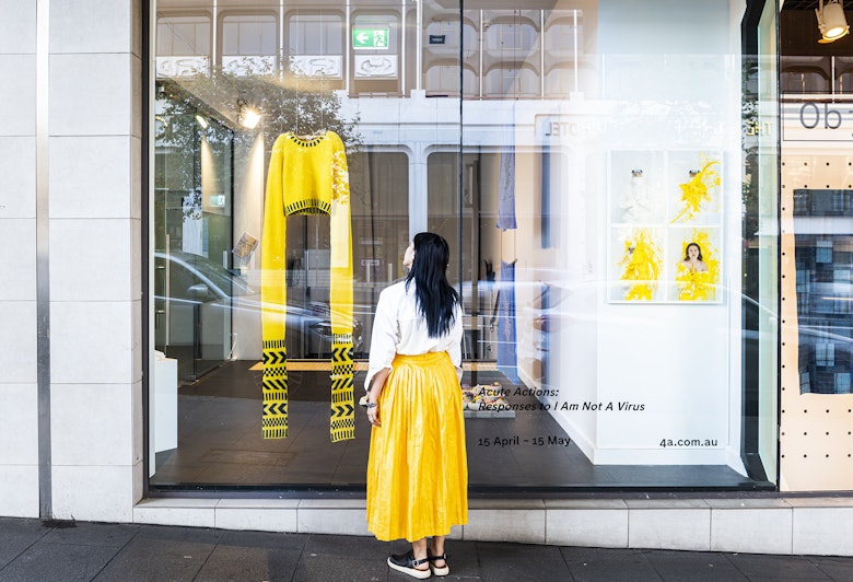 A woman with long black hair in a white blouse and yellow full-length skirt stands on a street outside a gallery glass front, looking at a yellow knitted sweater with sleeves knitted to be 1.5 metres long. The decal sign on the glass front reads 'Acute Actions: Responses to I Am Not A Virus. 15 April - 15 May'