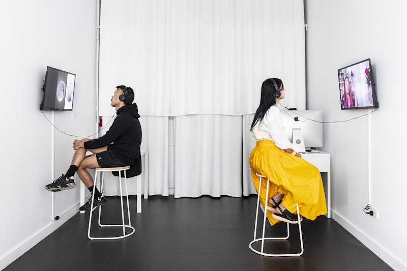 A male-presenting figure and a female-presenting figure sit back to back on white stools in a gallery space against a white curtain wall. The male figure is dressed in a black hoodie, black shorts and black socks and sneakers, with headphones over his ears as he watches a video work on a wall-mounted television screen. The female figure, dressed in a white blouse and a long cadmium yellow skirt, also has headphones over her ears and is watching a video work on a wall-mounted television screen.
