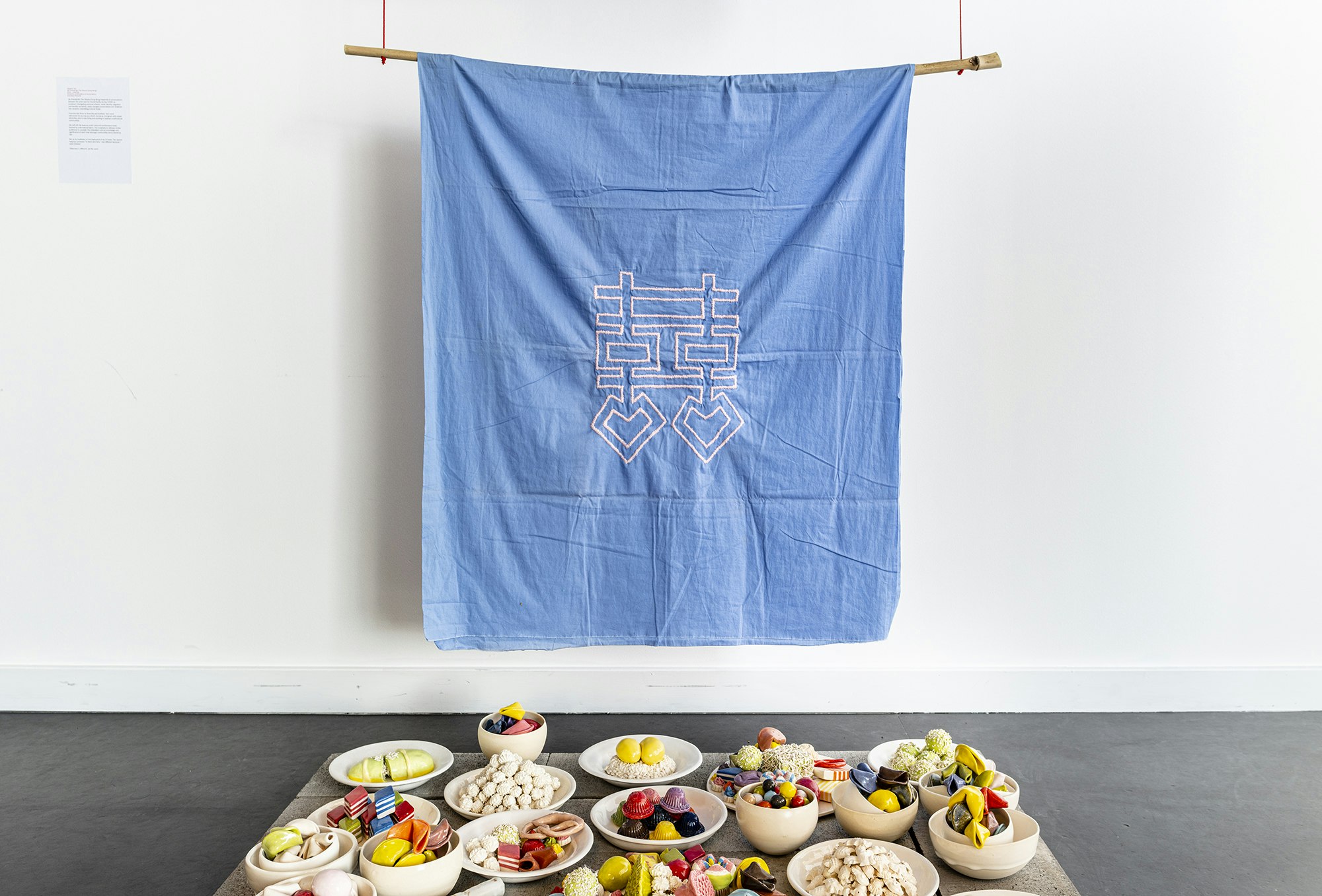 A colourful spread of earthenware sculptures imitating a spread of fruit, savoury snacks and East or Southeast Asian-inspired desserts. The spread features multicoloured fortune cookies, glutinous rice cakes, cakes rolled in shredded coconut and green, pink and red sticky rice cakes cut in the shapes of diamonds. Above this spread is a blue wall hanging embroidered with a fictional pink logogram.
