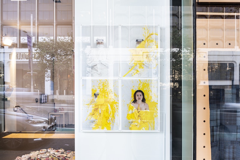 View from outside a gallery glass front of four photographic prints in white frames. The first shows a figure in a white hazmat suit and goggles standing against a blank white wall. Their palms are pressed together in prayer-like fashion front of their body, as a traditional Thai greeting. The second and third photograph show bright yellow paint being poured over the figure's head and splashing the wall behind them. The last photograph shows a female-presenting figure with the hazmat suit, now splashed with yellow paint, pulled down past her bare shoulders. Her palms are still pressed together as she looks at the camera.