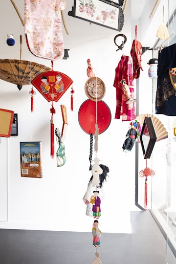 A series of objects hanging by a glass window, including a bright red Tang suit shirt, two pink cheongsam shirts, paper illustrations decorated with red Chinese knots, and a paper fan.