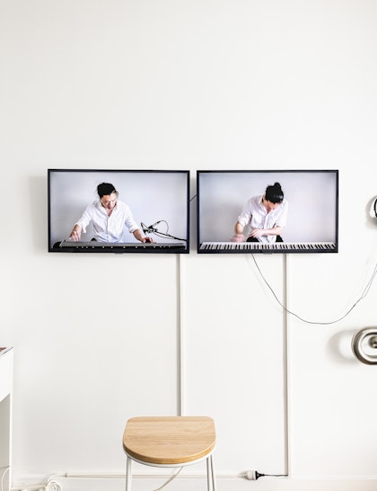 Two television screens next to each other on a gallery wall, showing an Asian male-presenting figure in a white button-up shirt playing on a portable keyboard. They have their black hair tied up in a bun.