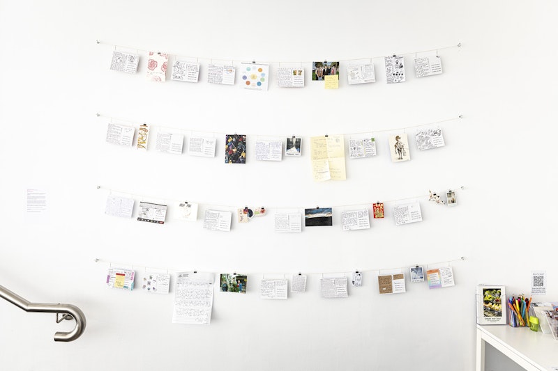 A white gallery wall with the corner of a white desk. On the wall are four hanging lines with postcards and letters pegged across them. On the table are stationery boxes holding coloured pens and postcards.