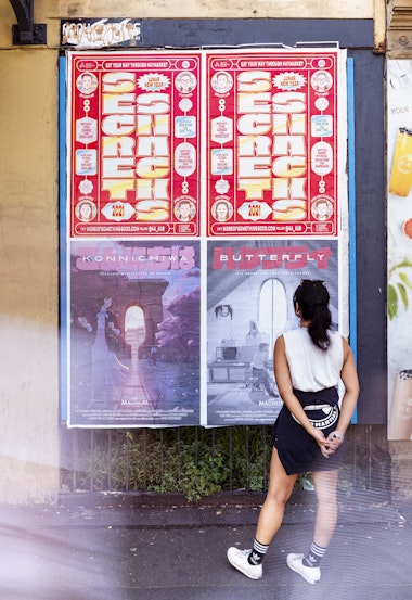  girl with a ponytail and a white singlet, shorts and sneakers looks up a wall at two red posters vertically printed with shiny gold letters that read 'SECRET SNACKS'. The posters are also printed with small colourful boxes of text and smiling cartoon faces of four Asian-Australians.