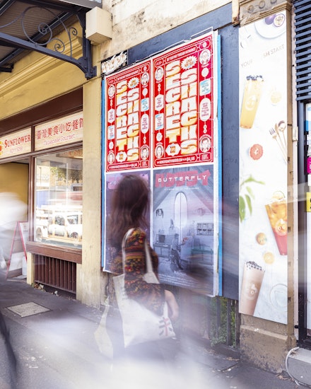 Two red posters printed with the words 'SECRET SNACKS' are stuck on a wall. A woman in a printed floral blouse carrying a white tote bag looks at the poster as she walks by.