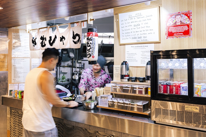 A man in a white singlet takes a bowl of ramen from a Japanese restaurant counter decorated with lanterns and a white noren curtain painted with kanji characters. A red square decal of a Thai-Australian man is stuck next to the window over a fridge with soft drinks. A whiteboard hanging near the window reads 'NO MSG'.