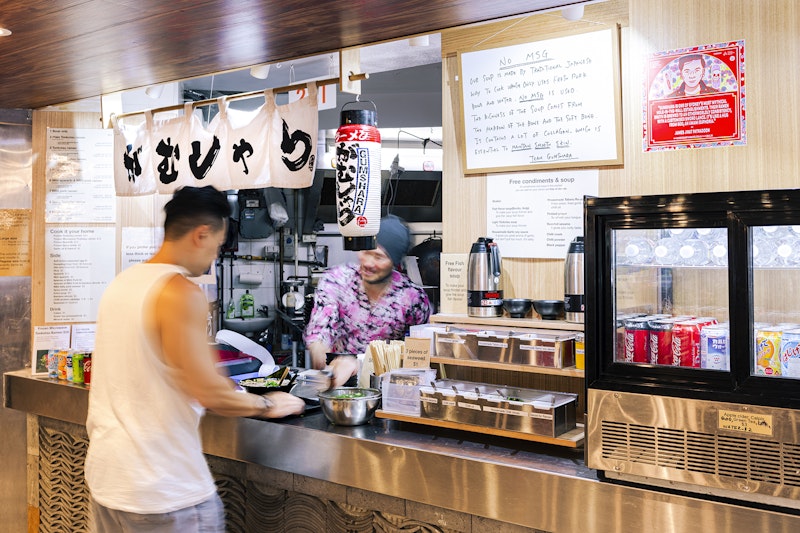 A man in a white singlet takes a bowl of ramen from a Japanese restaurant counter decorated with lanterns and a white noren curtain painted with kanji characters. A red square decal of a Thai-Australian man is stuck next to the window over a fridge with soft drinks. A whiteboard hanging near the window reads 'NO MSG'.