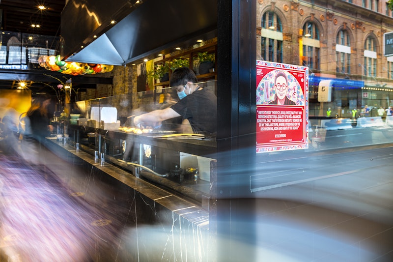 A red square decal of a Chinese-Australian woman with glasses is stuck on a restaurant door. Blurred figures pass in and out of the restaurant entrance while a chef in a black t-shirt and face mask works behind the kitchen window.