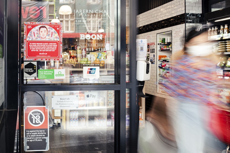 A red square decal of a Chinese-Australian man's face is stuck on a glass door outside a cafe and grocery store, as a blurred figure walks out.