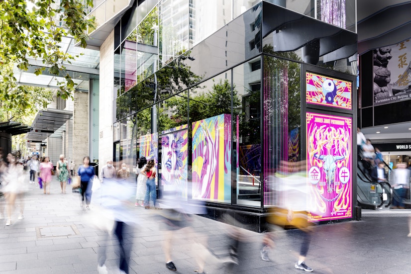 Pedestrians walk past a glass-fronted gallery with a neon mural of a woman's face with neon pink eyelids, green pupils and blue lips. A tall set of neon pink doors enclose the front of the gallery, painted with an ox skull surrounded by yellow flames and pink Chinese characters. Above the door is a cracked yin yang symbol surrounded by yellow flame with neon pink and blue rays shining from the yin yang.