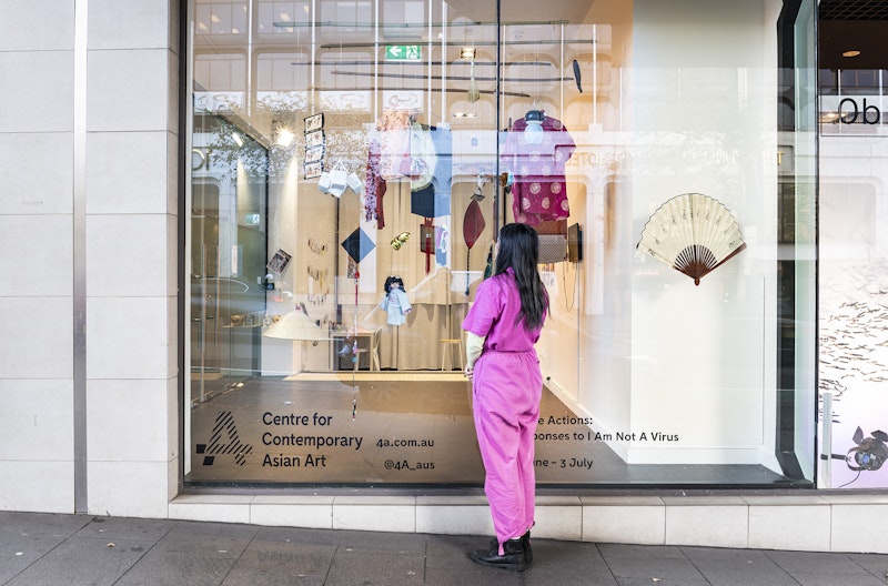 4A's gallery window with a paper fan, traditional Chinese silk garments, a doll and other textiles hanging from the ceiling. A woman with long black hair, wearing a fuchsia pink jumpsuit and black boots stands in front of the window, observing the installation.