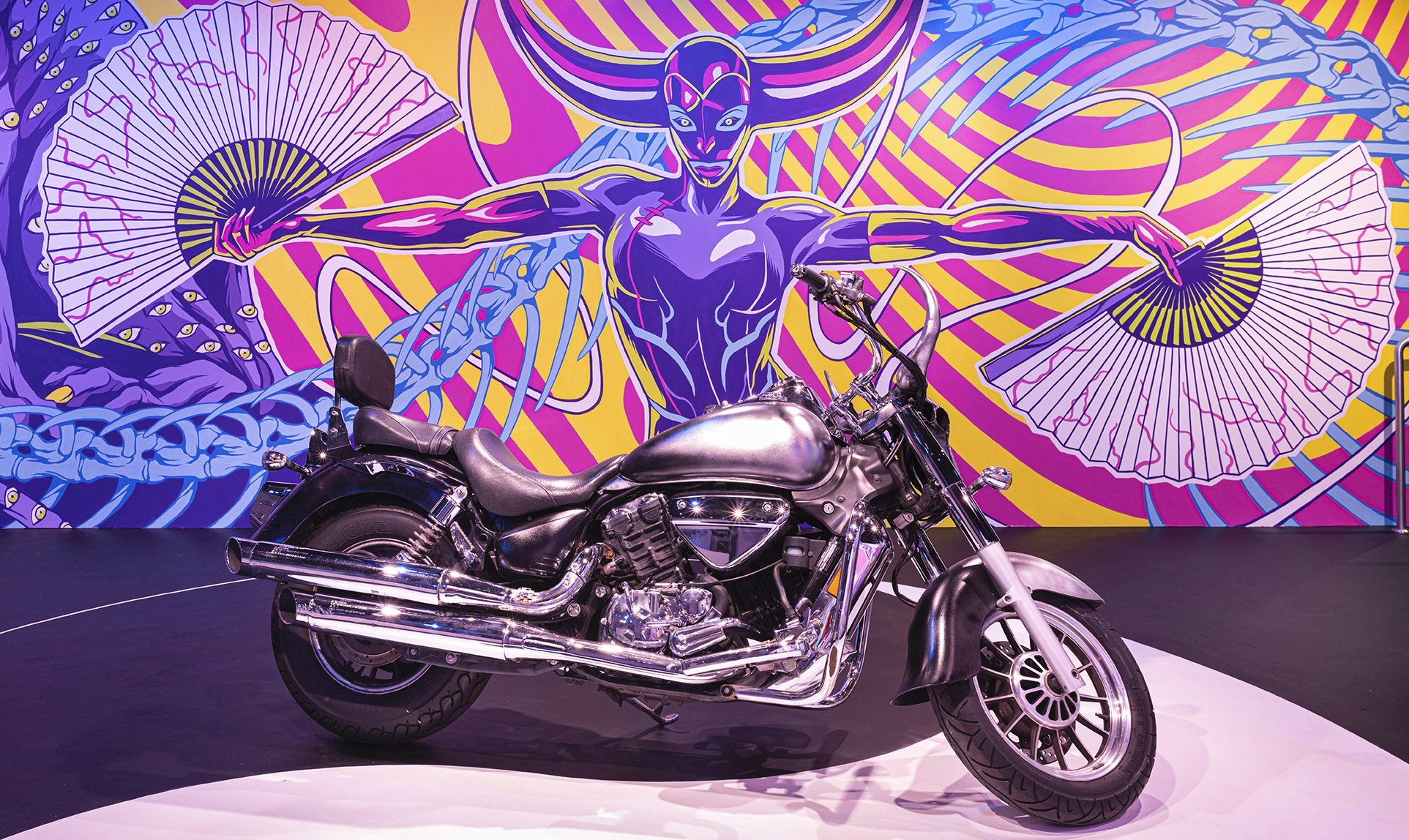 A chrome motorbike is parked in front of a mural of a feminised figure holding her arms out with an opened paper fan in each hand. Two large purple and pink ox horns protrude from her head, while she looks straight ahead with blue ringed eyes and green pupils. Behind her weaves a blue dragon's spine and a series of neon pink and yellow spirals.
