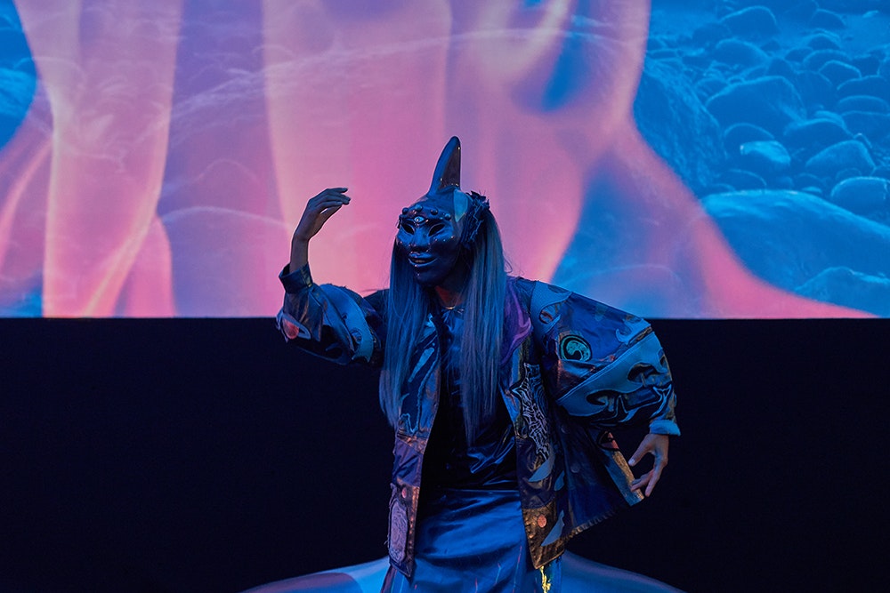 A costumed performer posing with one hand raised near their masked face, standing in front of a screen displaying red flame and blue rocks.