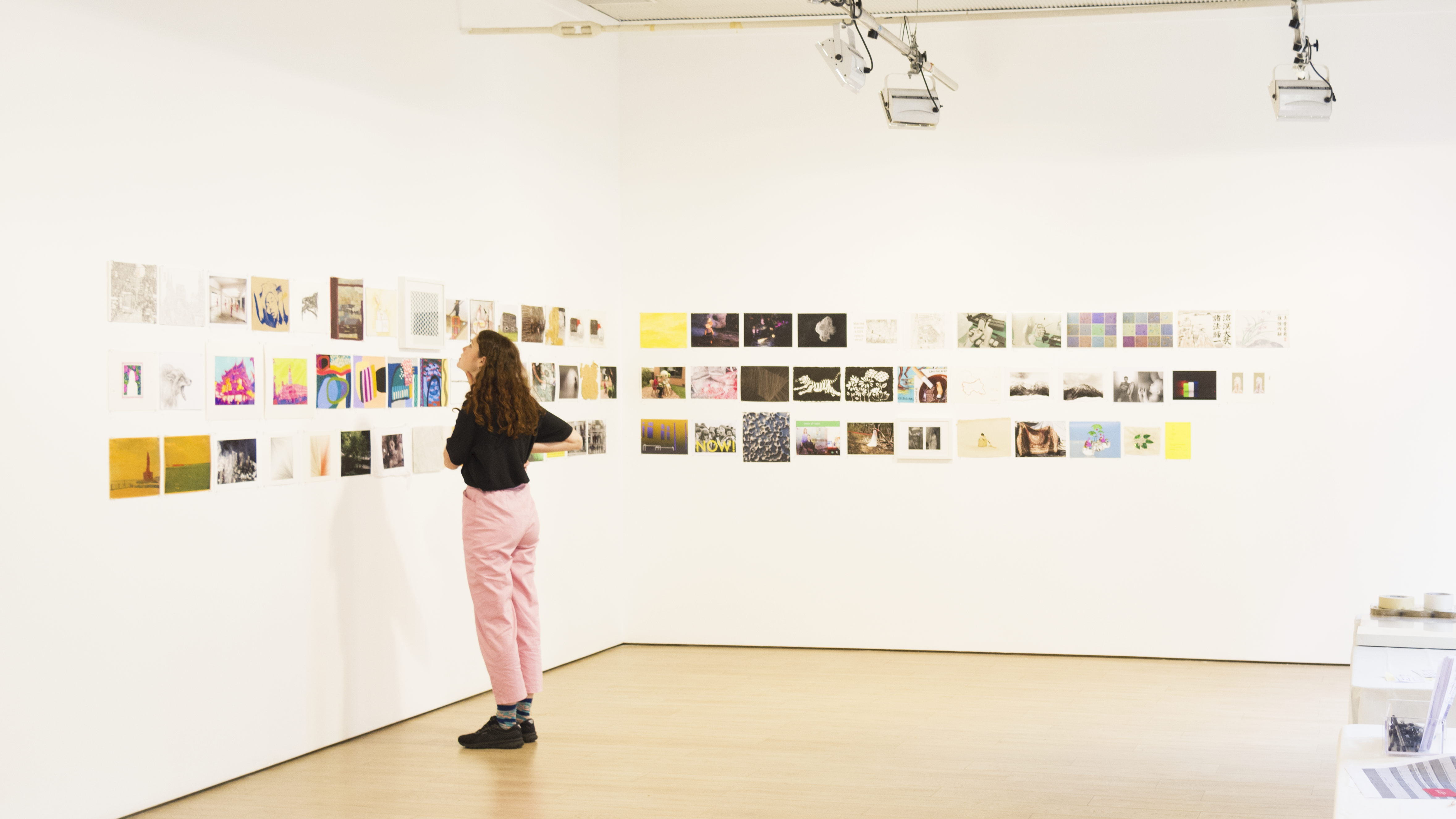 A woman with curly brown hair, wearing a black t-shirt, baby pink pants and black sneakers stands in an empty gallery, looking at a wall of A4-sized illustrations, paintings and designs.