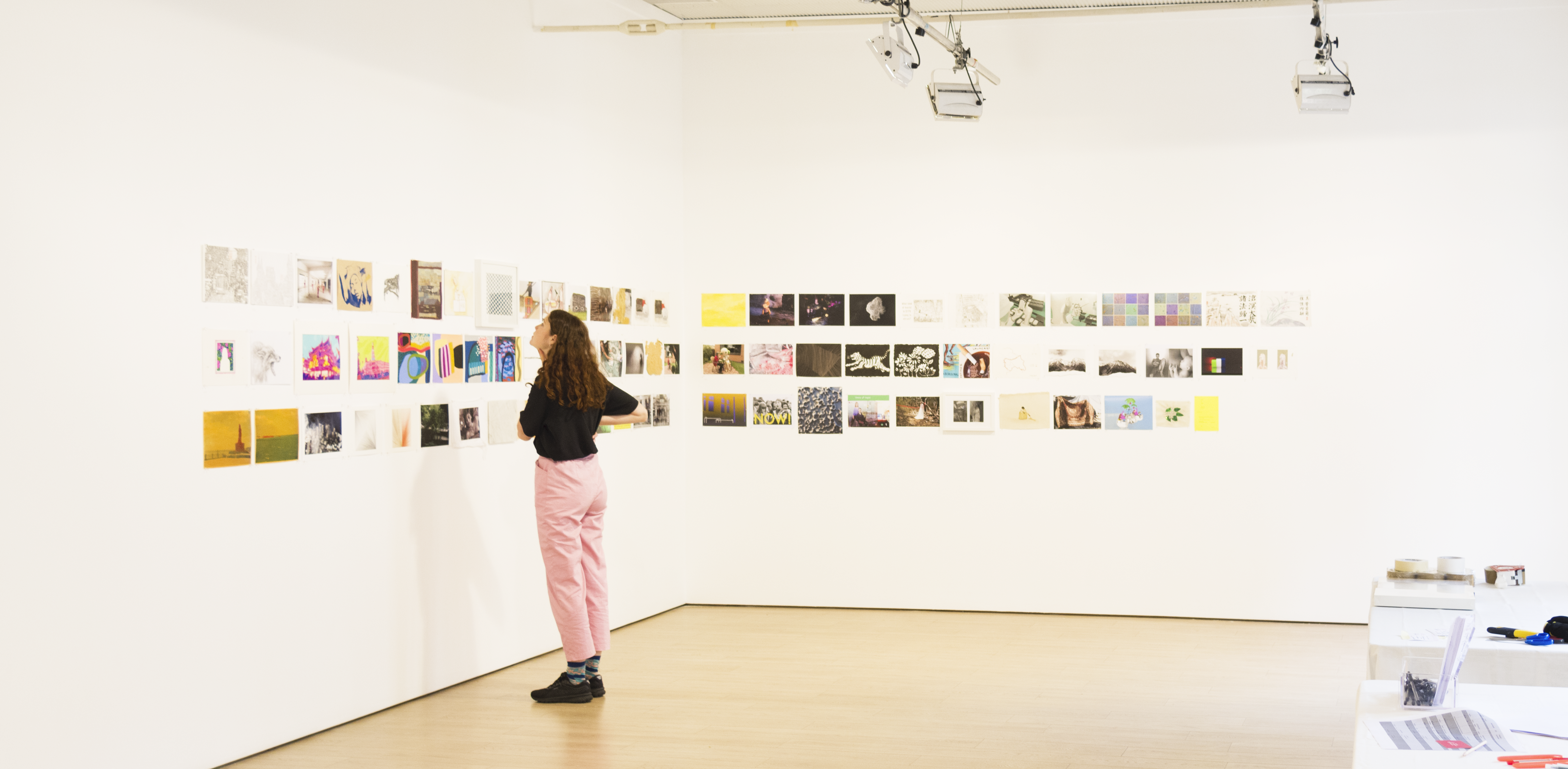 A woman with curly brown hair, wearing a black t-shirt, baby pink pants and black sneakers stands in an empty gallery, looking at a wall of A4-sized illustrations, paintings and designs.