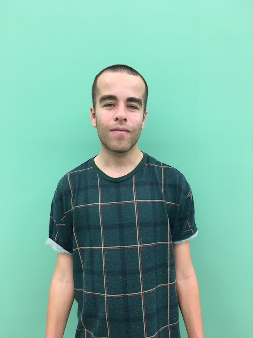 Rob stands in front of a green wall with a smile on his face and wears a green checkered t-shirt