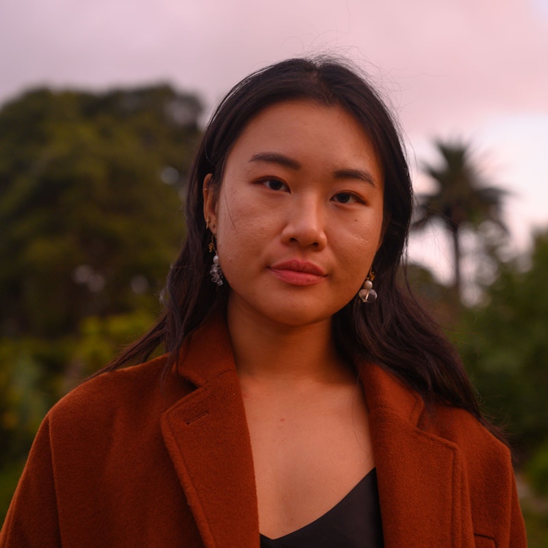 A tanned Vietnamese woman with medium length black hair slightly tucked behind her ears. She is wearing clear quartz earrings that resemble water droplets, standing against the faint backdrop of a garden at sunset. She is wearing a brown coat and a black silk camisole.